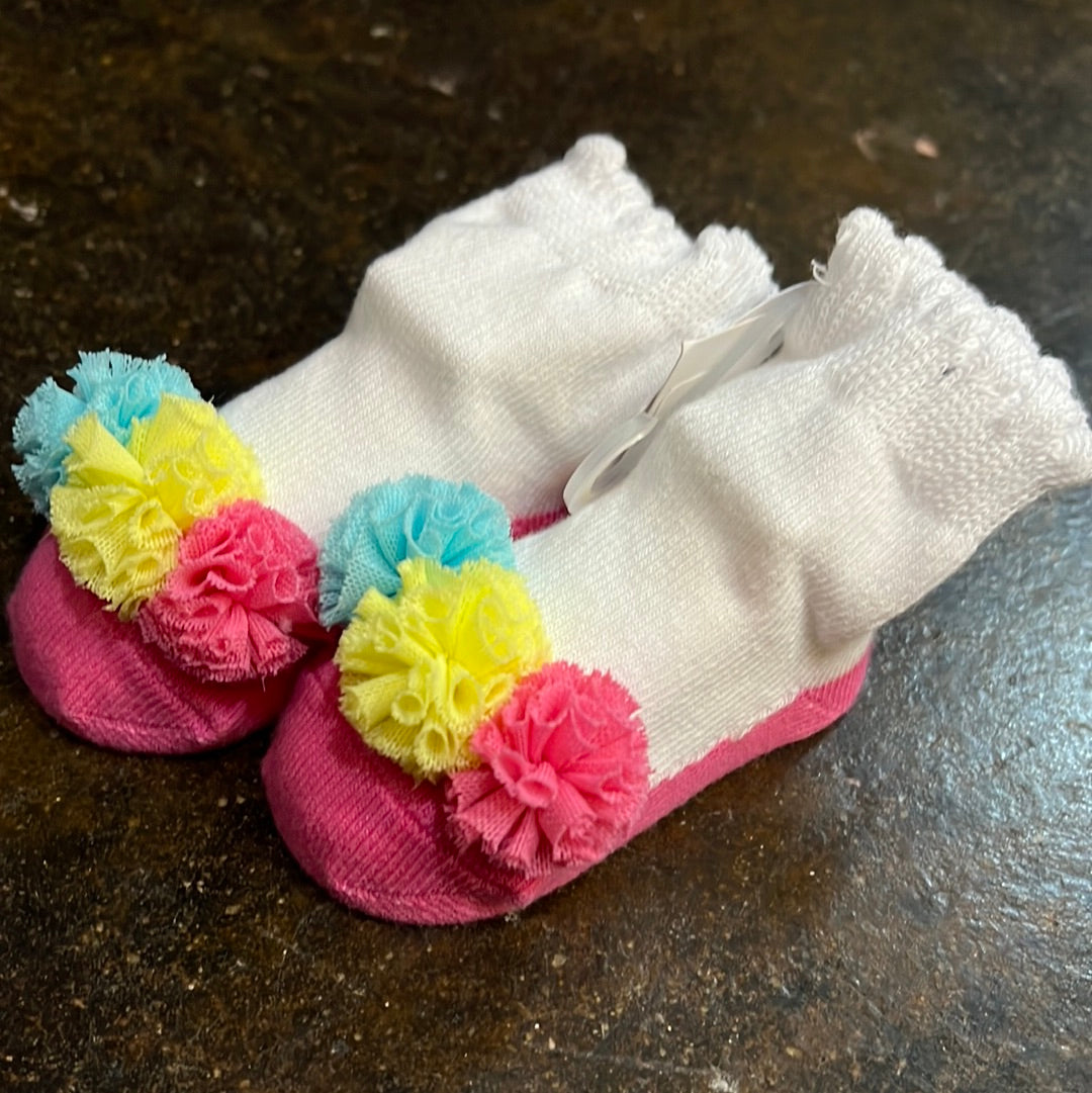 White baby socks with hot pink toes that have pink, yellow, and clue pom-poms on them.