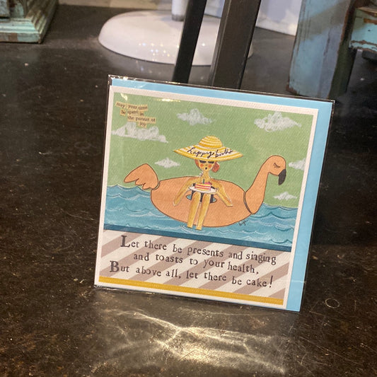 Card that featuring a woman floating in a pool flamingo. Says " Let there be presents and singing and toasts to your health. But above all, let there be cake!" Inside is blank.